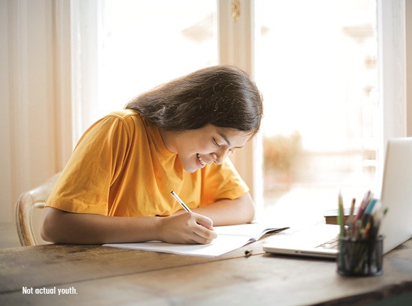 A young girl in a yellow shirt happily doing her homework at a table with a pen, paper, and a laptop